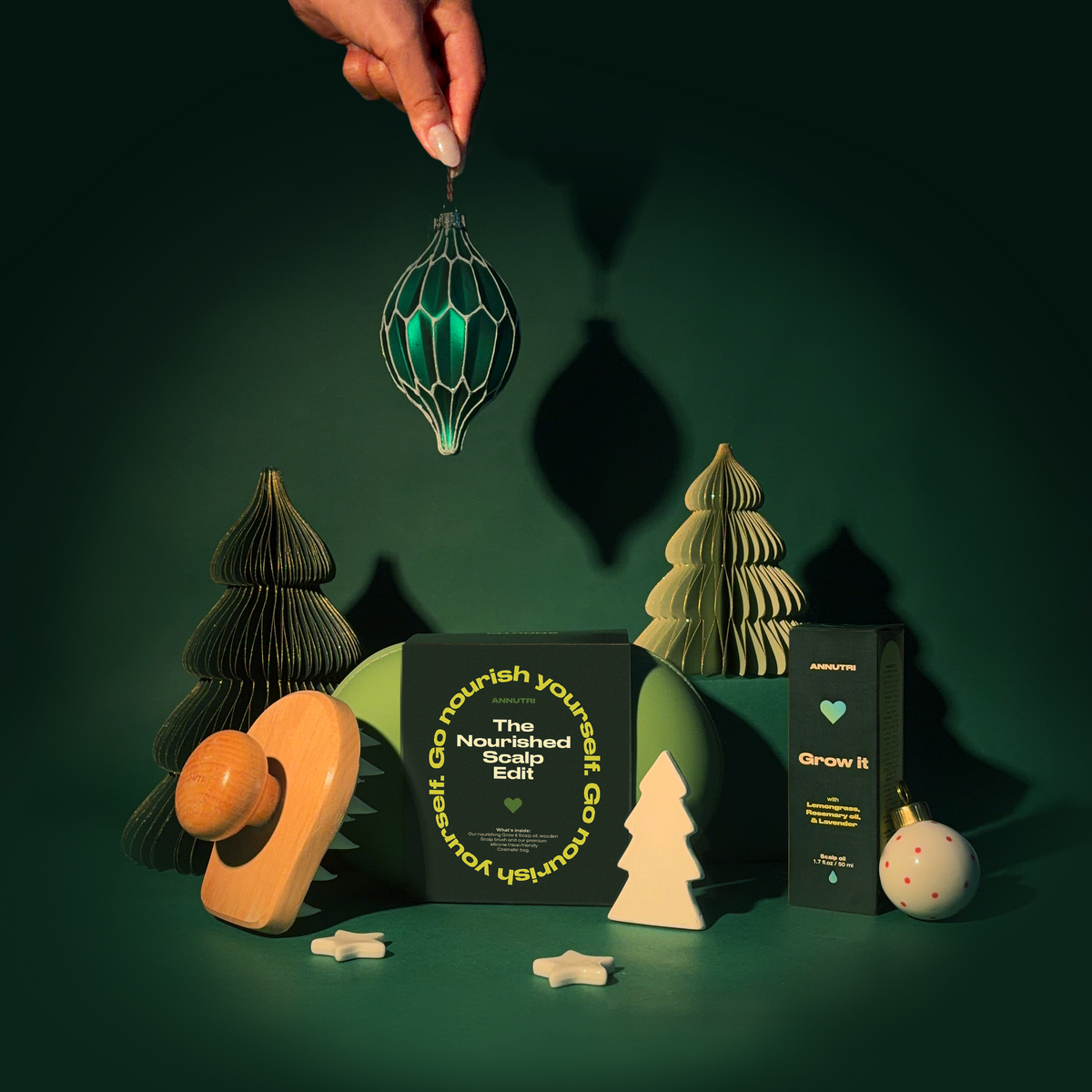 A hand holds a green hanging ornament above a set of Christmas-themed products, including a green box labeled 'The Christmas Tree Book', a dark green book, a beige paper tree, a wooden ornament, and small star and tree decorations, arranged on a dark green background.