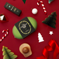 A styled flat-lay featuring a round green product with a black lid that reads 'The Christmas Gift Set' and 'Glow Up Yourself Go Glow Up Co.' surrounded by festive Christmas decorations on a deep red background.