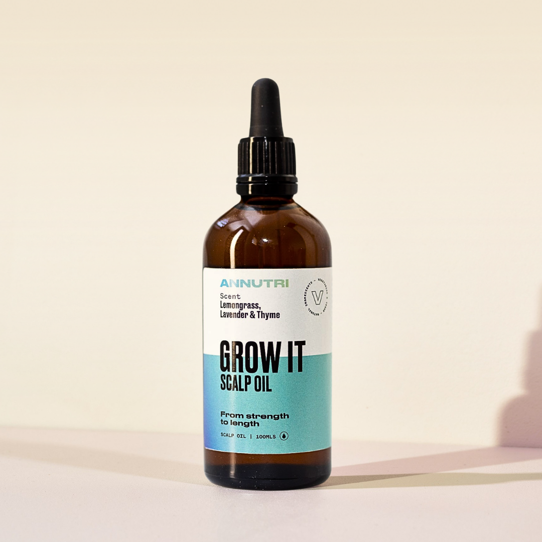 An amber glass dropper bottle of Annutri Grow It Scalp Oil with a black dropper cap and a white and teal label, set against a warm beige background.